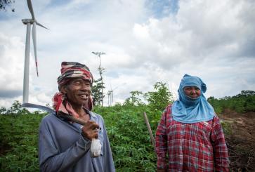 Two farmers stand in their field in Thailand, in front of wind turbines.