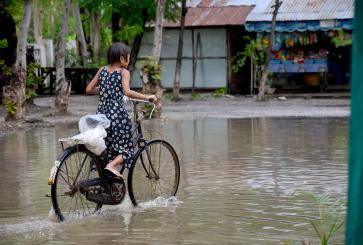A girl wearing a long sundress pedals a bicycle down a flooded road..